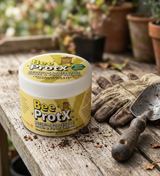 A jar of BeeProtX Beeswax Protective Hand & Foot Cream sits on a rustic wooden table outdoors, styled with gardening gloves and a trowel. The background shows blurred potted plants.