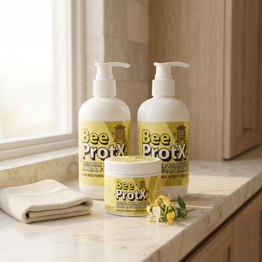 Three BeeProtX hand and foot care products arranged on a marble countertop near a window: two large white pump bottles and one smaller white tub with yellow labels, plus a folded beige towel and small yellow flowers.