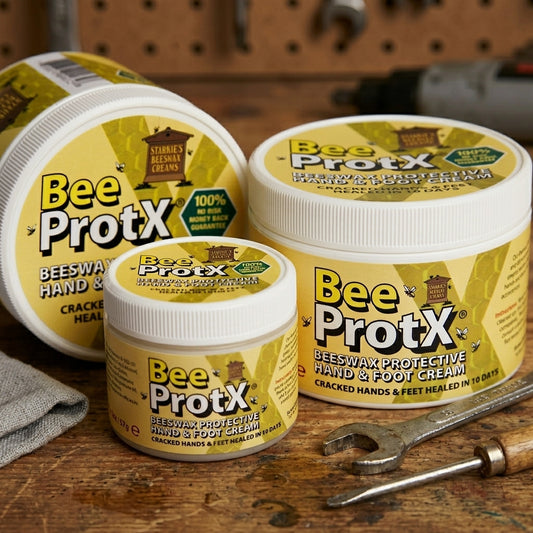 Three tubs of BeeProtX Beeswax Protective Hand & Foot Cream arranged on a rustic wooden workbench with a wrench and cloth, showing the product packaging in a workshop-like setting.