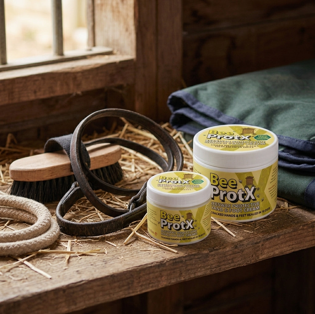 Two tubs of BeeProtX Barrier Hand & Foot Cream placed on a rustic wooden stable bench with hay, horse grooming tools, and a folded green blanket nearby.