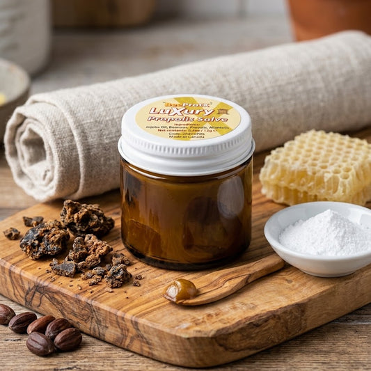 A jar of luxury BeeProtX Propolis salve displayed on a wooden cutting board with its ingredients, including raw honeycomb, white wax, coffee beans, and a wooden spoon, alongside a rolled cloth and a small bowl of powder.