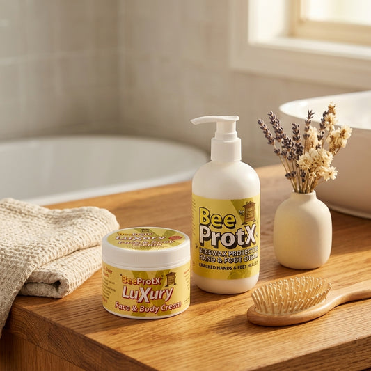 BeeProtX Luxury Face & Body Cream tub and Hand & Foot Cream pump bottle displayed on a wooden bathroom counter next to a towel, hairbrush, and vase with dried lavender.